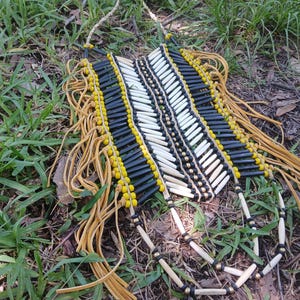 May include: A breastplate necklace featuring rows of black and white cylindrical beads arranged in a chevron pattern, accented with yellow and metallic spacer beads, finished with long tan leather fringe strands hanging from the sides.