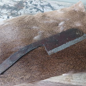 May include: A rusty, hand-forged knife with a curved handle and a sharp blade. The knife is lying on a brown fur surface.