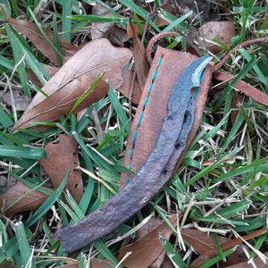 May include: A small, rusty, handmade knife with a curved blade, in a brown leather sheath with blue stitching. The knife is lying on a bed of green grass and brown leaves.