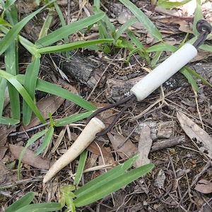 May include: A white cylindrical bead and a tapered, pointed antler tip connected by a dark brown leather cord. The cord forms a loop, securing the two components together in a simple arrangement with a knot at the end of the leather cord.