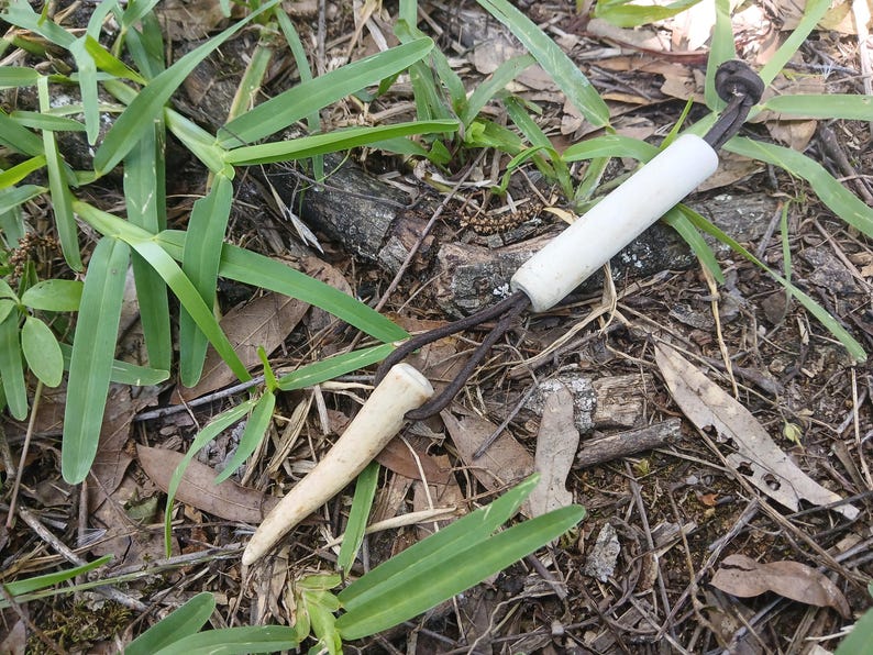 May include: A white cylindrical bead and a tapered, pointed antler tip connected by a dark brown leather cord. The cord forms a loop, securing the two components together in a simple arrangement with a knot at the end of the leather cord.