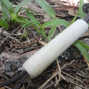 May include: A cylindrical white handle with a smooth finish, featuring a dark brown leather cord threaded through the centre. The cord exits both ends of the handle, with one end tied into a simple knot, creating a functional grip or pull tool.