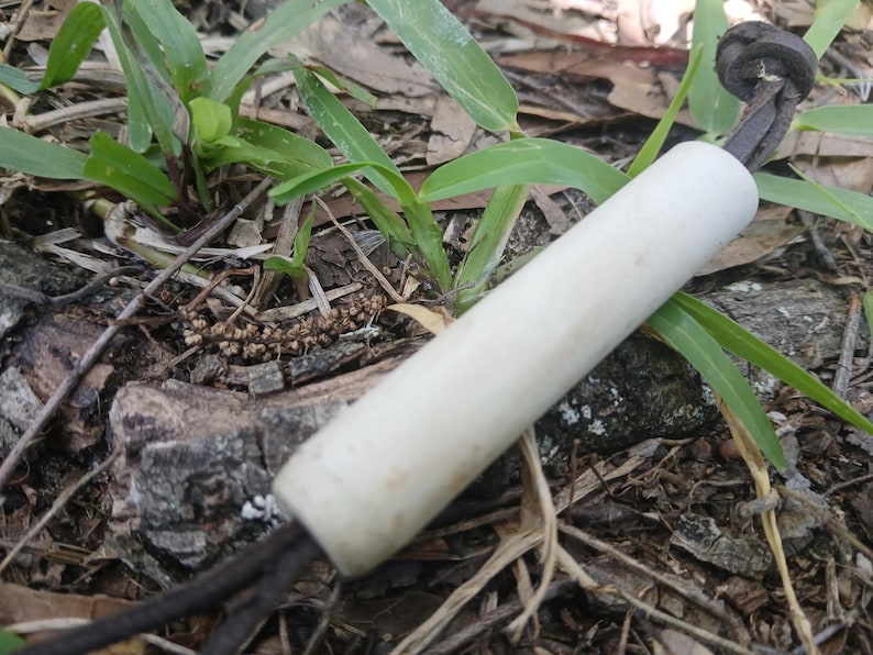 May include: A cylindrical white handle with a smooth finish, featuring a dark brown leather cord threaded through the centre. The cord exits both ends of the handle, with one end tied into a simple knot, creating a functional grip or pull tool.