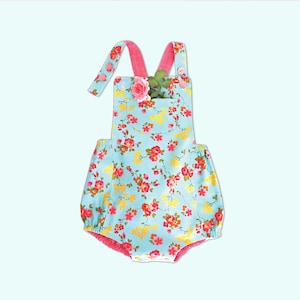 May include: A light blue baby romper with a floral print featuring pink, yellow, and red flowers. The romper has pink straps and a single button on each strap. The romper has a pocket on the front with a pink flower tucked into it.