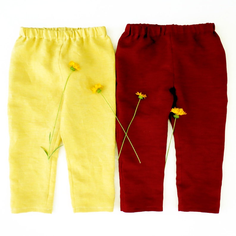 Puede incluir: Dos pantalones, uno amarillo y otro burdeos, est&aacute;n colocados sobre una superficie blanca. Ambos pantalones est&aacute;n hechos de una tela suave, similar al lino.