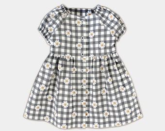 Puede incluir: Un vestido de cuadros vichy blanco y negro con estampado de margaritas blancas y mangas cortas. El vestido tiene un cierre de botones en la parte delantera.