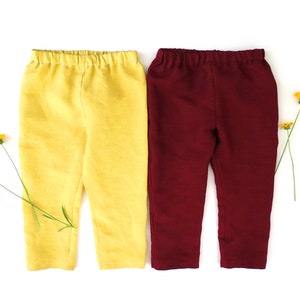 Puede incluir: Dos pares de pantalones para ni&ntilde;os, uno amarillo y otro burdeos. Los pantalones est&aacute;n colocados sobre un fondo blanco.