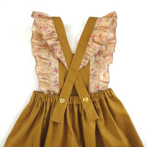 May include: A mustard yellow overall dress with a gathered skirt and criss-cross straps. The straps are trimmed with a floral print fabric with ruffles.