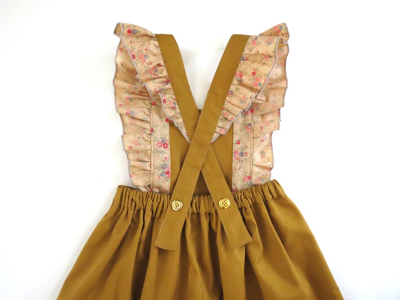 May include: A mustard yellow overall dress with a gathered skirt and criss-cross straps. The straps are trimmed with a floral print fabric with ruffles.