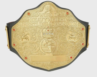 Puede incluir: Un cinturón de campeonato de lucha libre dorado con las palabras "WORLD HEAVYWEIGHT WRESTLING CHAMPION" grabadas. El cinturón presenta diseños intrincados, una corona y joyas rojas. El fondo es gris claro.
