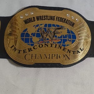 Puede incluir: Un cinturón de campeonato Intercontinental dorado y negro. El cinturón presenta el texto "WORLD WRESTLING FEDERATION" e "INTERCONTINENTAL CHAMPION" en letras doradas. La placa central tiene un diseño de mapa del mundo azul con el logotipo de la WWF.