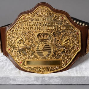 Puede incluir: Un cinturón de campeonato de lucha libre de cuero marrón con una gran placa central de metal dorado ornamentada. La placa está grabada con el texto WORLD HEAVYWEIGHT WRESTLING CHAMPION, una corona, un globo terráqueo y volutas intrincadas con detalles rojos.