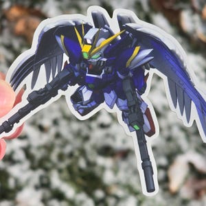 May include: A sticker featuring a detailed illustration of a Gundam Wing Zero robot. The robot is primarily blue, white, and yellow, with large, feathered wings and two rifles. The sticker is held against a blurred, natural background.