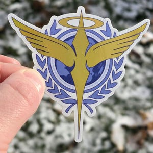 May include: A sticker featuring a gold emblem with a stylized bird design, wings outstretched, set against a blue and white background. The emblem includes a halo and a globe, surrounded by a laurel wreath. The sticker is held against a blurred, snowy backdrop.
