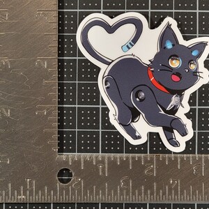 May include: A black and white cartoon sticker of a robot cat with a red collar and a heart-shaped tail. The cat has blue eyes and a surprised expression.