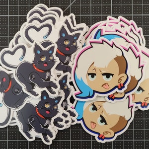May include: A set of stickers featuring a cartoon character with blue hair and a tongue sticking out, and a black cat with a heart-shaped tail. The stickers are printed on white paper and have a glossy finish.