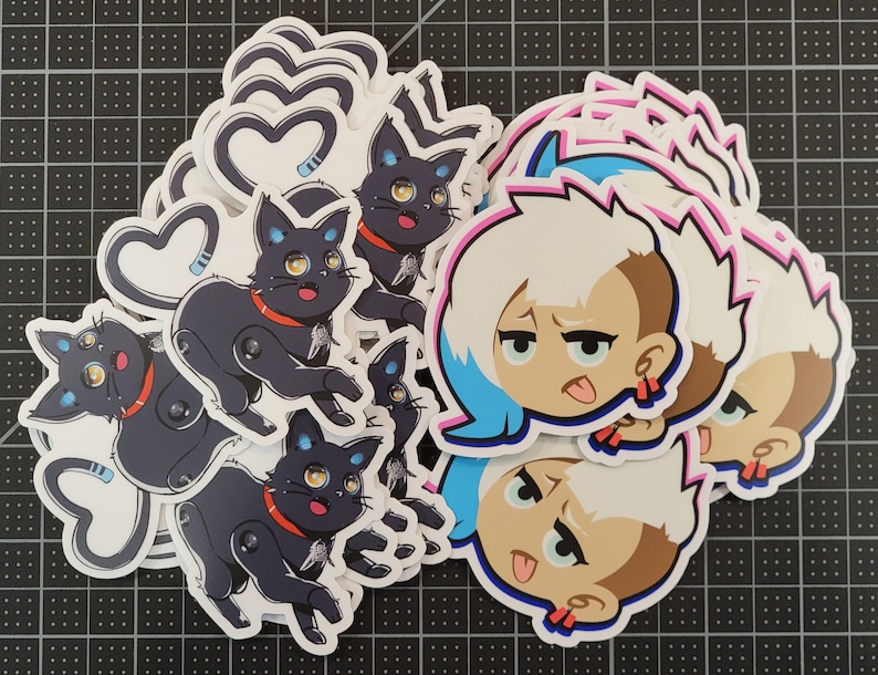 May include: A set of stickers featuring a cartoon character with blue hair and a tongue sticking out, and a black cat with a heart-shaped tail. The stickers are printed on white paper and have a glossy finish.