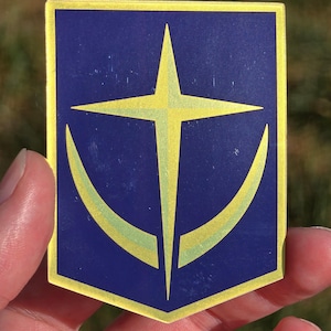 以下が含まれることがあります： 金と青の盾型のステッカーで、金の星と三日月のデザインが施されています。