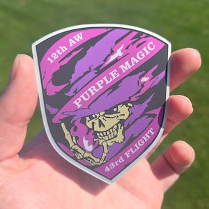 Può includere: Un adesivo a forma di scudo con una grafica a teschio e segni di artigli viola e rosa. Il testo riporta 12th AW, PURPLE MAGIC e 43rd FLIGHT. Il design presenta uno sfondo scuro con un teschio beige e fiamme di colore viola.