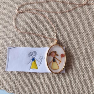 Op de afbeelding: Een goudkleurige ketting met een ovale hanger met een kindertekening. De tekening toont een stokfiguur met een bloem. Een bijpassende tekening staat op een klein stukje papier naast de hanger.
