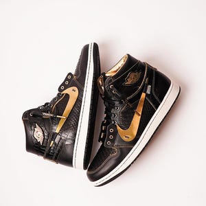 Peut inclure: Paire de baskets montantes noires et dorées. Les baskets présentent un swoosh doré, des accents dorés et une semelle blanche. Le mot "PANDA" est visible sur une chaussure. Les chaussures sont présentées sur fond blanc.