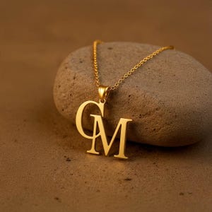 May include: A gold-colored necklace with the letters "GM" as a pendant. The pendant is suspended from a delicate chain. The necklace is displayed on a textured, light brown stone, set against a neutral background.