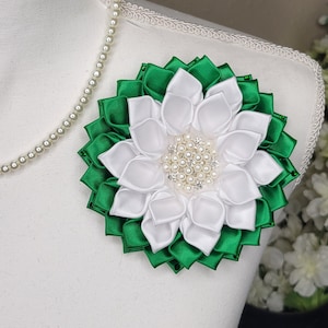 May include: A floral brooch in white and green satin ribbon, with a pearl and rhinestone center. The brooch is attached to a white mannequin. A pearl necklace is draped around the mannequin's neck.