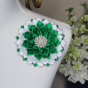 May include: A decorative floral brooch made of layered green and white satin ribbons. The brooch is centered with a cluster of faux pearls and rhinestones. The brooch is pinned to a white mannequin.