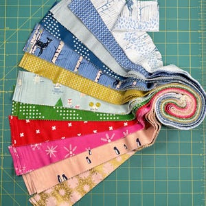 Peut inclure: Une collection de bandes de tissu de différentes couleurs et motifs, dont des cerfs, des arbres et des flocons de neige. Les bandes sont disposées en éventail, avec un paquet roulé à l'extrémité. Les tissus sont sur un tapis de coupe vert.