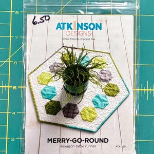 May include: A pattern for a hexagon table runner called "Merry-Go-Round" by Atkinson Designs. The pattern features a white background with green, blue, and gray hexagons. The pattern is $6.50 and is labeled ATK-169.