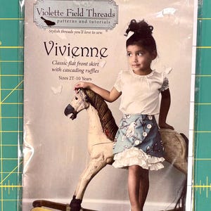 May include: A sewing pattern for a girl's skirt called "Vivienne" by Violette Field Threads. The pattern features a classic flat front skirt with cascading ruffles. The pattern is for sizes 2T-10 years.