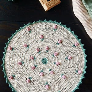 Op de afbeelding: Een crèmekleurige, ronde gehaakte doily met een groene geschulpte rand en kleine roze bloemaccenten. De doily is handgemaakt en heeft een gedetailleerd, getextureerd ontwerp. Garen en koekjes staan op de achtergrond.