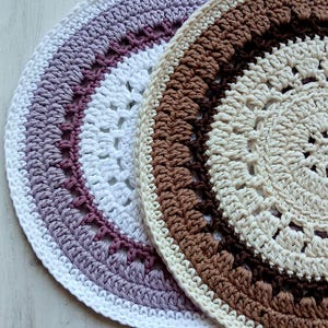 以下が含まれることがあります： 手作りのかぎ針編みコースター2枚。1枚は白、紫、ボルドーの色使い。もう1枚はクリーム、茶色、濃い茶色の色使い。どちらのコースターも円形で、レースのようなオープンデザインです。