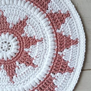 May include: A circular, hand-crocheted rug with a repeating pattern in white and dusky pink. The design includes a central floral motif and concentric rings of the two colours. The edge is finished with a white border.
