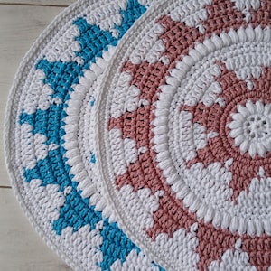 Op de afbeelding: Twee handgemaakte gehaakte placemats. Eén is wit met turkooizen accenten en de andere is wit met stoffige roze accenten. De ronde matten hebben een zich herhalend patroon. De placemats liggen op een licht houten oppervlak.