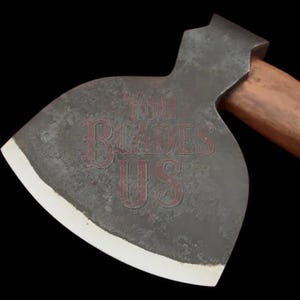 Könnte beinhalten: Nahaufnahme einer Vintage-Axt mit einem dunkelgrauen Kopf und einem Holzgriff. Der Axtkopf hat eine weiße Kante und der Text "YOU BLADES US" ist auf der Klinge sichtbar. Der Hintergrund ist schwarz.