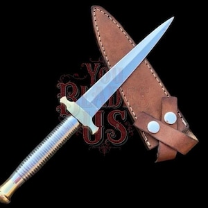 Puede incluir: Una daga plateada con una hoja de doble filo y un mango plateado acanalado. La guarda y el pomo son dorados. La daga descansa sobre una funda de cuero marrón con dos cierres de presión. El fondo es negro.