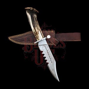 Pode incluir: Uma faca de caça com uma lâmina serrilhada e um cabo de chifre. A faca está em uma bainha de couro marrom com uma presilha para cinto. A lâmina tem texto gravado. O fundo é preto.