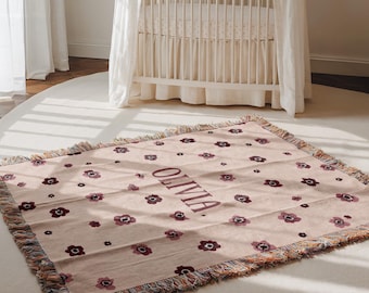 May include: A light pink woven blanket with a floral pattern and the name "OLIVIA" in dark brown lettering. The blanket has a fringed edge and is placed on a white rug in front of a crib.