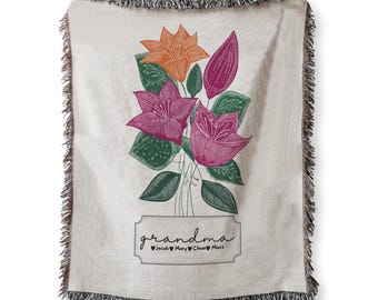May include: Cream-colored throw blanket with a floral design in orange, pink, and purple. The word "grandma" is written in cursive below the flowers, with names below. The blanket has a fringed edge.