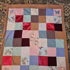 Peut inclure: Une courtepointe patchwork avec une variété de tissus roses, bleus, bruns et bordeaux. Certains carrés présentent des motifs floraux, tandis que d'autres ont des couleurs unies ou de petites broderies. La courtepointe a une bordure marron et un motif brodé central.