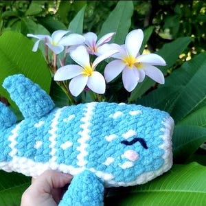 May include: A handmade, crocheted whale plushie in shades of blue and white, with a smiling face. The plush toy is set against a backdrop of green leaves and white flowers, creating a soft, whimsical aesthetic.