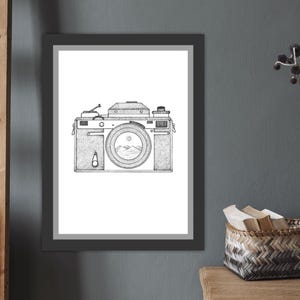 May include: Black and white framed art print featuring a detailed illustration of a vintage camera. The camera's lens showcases a mountain landscape. The artwork is displayed on a gray wall, with a wooden ladder and decorative items in the background.