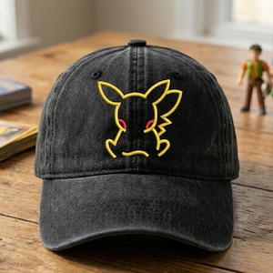 Könnte beinhalten: Schwarze Baseballkappe mit gelbem Pikachu-Stickmuster. Die Kappe hat einen gebogenen Schirm und einen Used-Look. Das Pikachu-Design hat gelbe Umrisse und rote Augen. Die Kappe liegt auf einer Holzoberfläche.