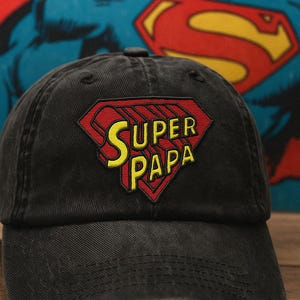 May include: Black baseball cap with a red and yellow "Super Papa" logo. The logo is in a diamond shape, reminiscent of a superhero emblem. The cap is made of a durable fabric and has a curved brim.