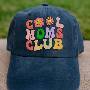 May include: Navy blue baseball cap with the embroidered text "COOL MOMS CLUB" in colorful, retro-style lettering. The design includes a smiling flower and a star. A casual, everyday accessory.