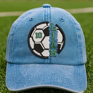 K&ouml;nnte beinhalten: Hellblaue Jeans-Baseballkappe mit einem Fu&szlig;ball-Design. Der Fu&szlig;ball ist schwarz und wei&szlig; mit dem Namen "LIAM" in gr&uuml;nen Buchstaben. Die Nummer "10" ist ebenfalls gr&uuml;n. Die Kappe befindet sich auf einem gr&uuml;nen Feld.