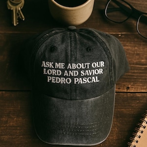 May include: A dark grey baseball cap with white embroidered text reading "ASK ME ABOUT OUR LORD AND SAVIOR PEDRO PASCAL." The cap sits on a wooden surface with a coffee mug, keys, glasses, and a notebook.