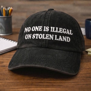 May include: Black baseball cap with white embroidered text reading "NO ONE IS ILLEGAL ON STOLEN LAND". The cap sits on a wooden surface, with stationery and a mug visible in the background.