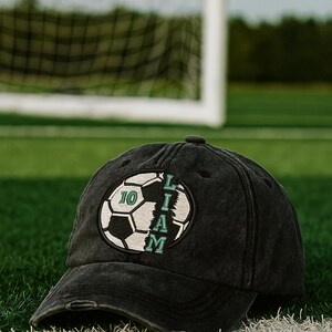 K&ouml;nnte beinhalten: Schwarze Baseballkappe mit Used-Look, mit einem Fu&szlig;ball-Design mit der Nummer 10 und dem Namen "LIAM" in Gr&uuml;n und Wei&szlig;. Die Kappe liegt auf einem gr&uuml;nen Feld mit einer wei&szlig;en Linie, mit einem Fu&szlig;balltor im Hintergrund.